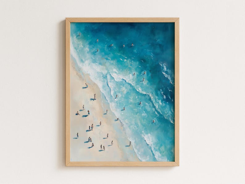 May include: A framed art print depicts a beach scene from an aerial perspective. The artwork features a sandy shore, turquoise waves, and numerous tiny figures of people. The frame is made of light-colored wood.