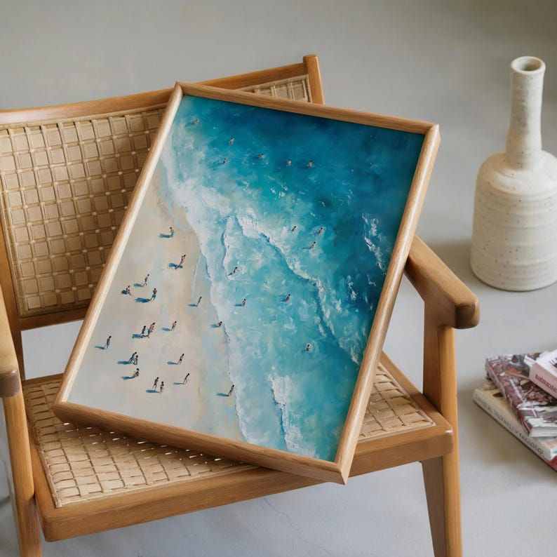 May include: A framed art print depicting a beach scene with turquoise water and a sandy shore. The artwork features small figures of people and is set in a light wood frame. The print is displayed on a wooden chair with woven seat.