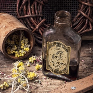 Pu&ograve; includere: Una bottiglia di vetro marrone con l'etichetta "Unicorn Blood" e un'illustrazione di un unicorno. L'etichetta recita anche "Mystic Apothecary". Erbe secche e un contenitore di legno su una superficie di legno, creando un'estetica da erboristeria mistica.