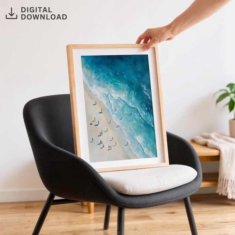 May include: A framed art print featuring an aerial view of a beach with turquoise water and white waves. The print is in a light wood frame and is labeled "DIGITAL DOWNLOAD". The print is displayed on a black chair with a white cushion.
