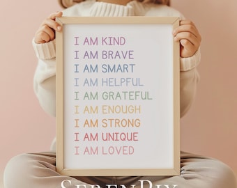 May include: A child holds a white framed sign with the text "I am kind, I am brave, I am smart, I am helpful, I am grateful, I am enough, I am strong, I am unique, I am loved." The sign has a light wood frame and the text is in rainbow colors. The child is wearing a white sweater and beige pants. The background is a light pink wall.