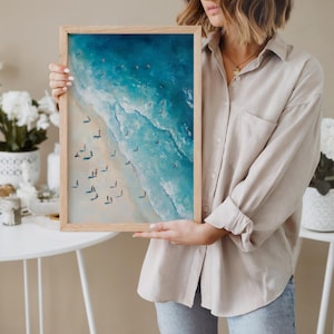May include: A framed art print depicting an aerial view of a beach scene. The artwork features a vibrant blue ocean with white waves crashing onto a sandy shore. Small figures of people are scattered along the beach and in the water.