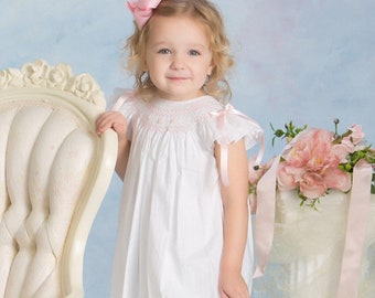 May include: A young girl wearing a white smocked dress with pink ribbon accents and a pink bow in her hair. She is standing next to a white chair and a vase of pink flowers.