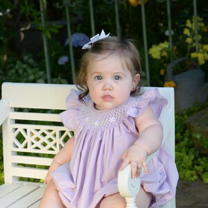 May include: A young child wearing a lavender smocked dress with a white bow in their hair sits on a white wooden bench.