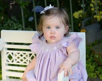 May include: A young child wearing a lavender smocked dress with a white bow in their hair sits on a white wooden bench.