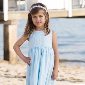 Pode incluir: Uma menina pequena com um vestido azul claro sem mangas, bordado branco e acabamento em renda. Ela está em pé em uma praia de areia e está usando uma faixa de flores brancas.