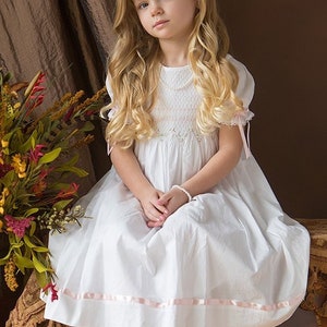 May include: A white dress featuring short puff sleeves with pink ribbon ties, a smocked bodice with delicate embroidery, and a hemline accented by a horizontal pink ribbon. The dress has a round neckline and a full, flowing skirt silhouette.