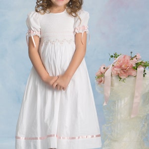 May include: A white dress featuring short puff sleeves with pink ribbon ties, a smocked bodice with delicate pink floral embroidery, and a pink ribbon trim along the hem. The dress has a round neckline and a full skirt that falls below the knees.