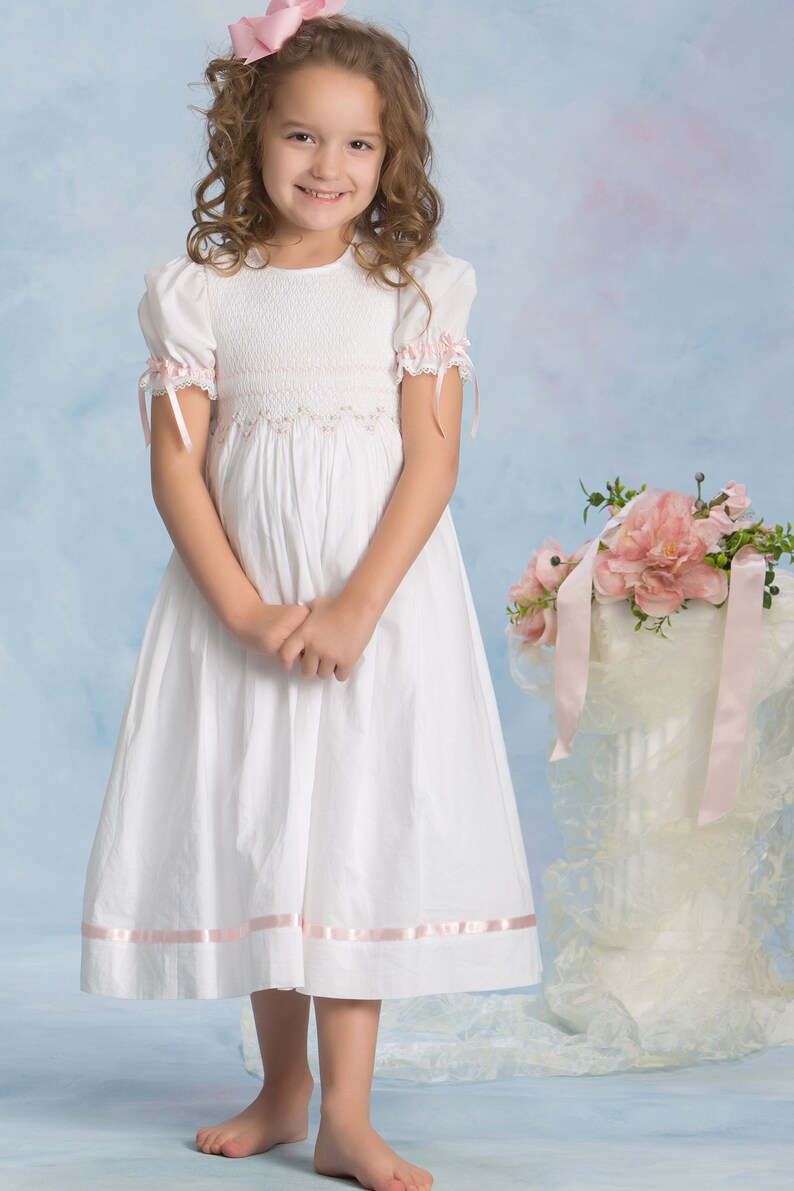 May include: A white dress featuring short puff sleeves with pink ribbon ties, a smocked bodice with delicate pink floral embroidery, and a pink ribbon trim along the hem. The dress has a round neckline and a full skirt that falls below the knees.