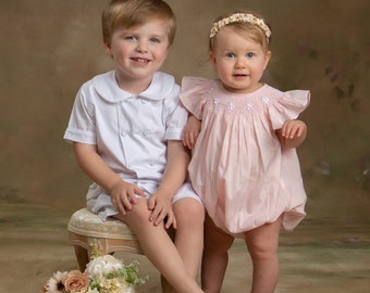May include: A young boy wearing a white button-down shirt and shorts sits on a stool with a bouquet of flowers in front of him. A young girl wearing a pink smocked dress with a floral headband stands next to him.