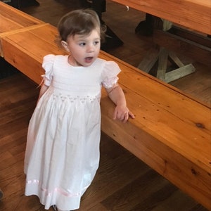 May include: A white floor-length dress with short puffed sleeves, featuring delicate smocking at the bodice and waist with subtle pink floral embroidery. The hem is accented with a thin pink ribbon trim, and the dress is paired with white sandals.