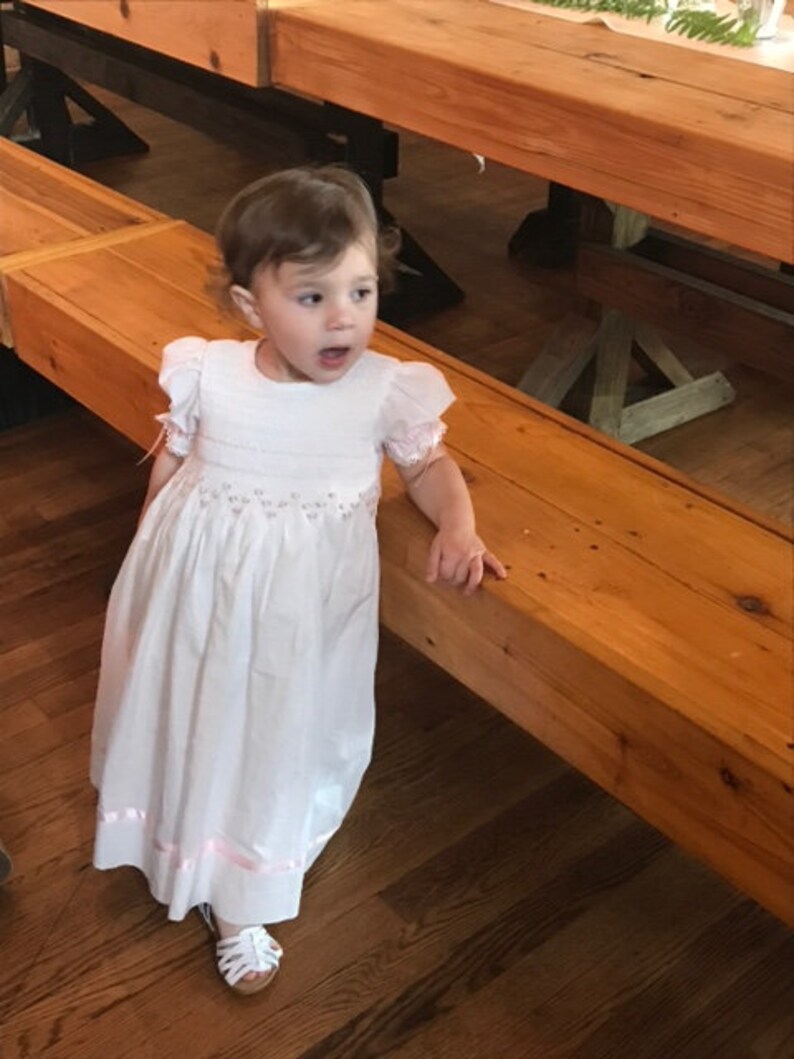 May include: A white floor-length dress with short puffed sleeves, featuring delicate smocking at the bodice and waist with subtle pink floral embroidery. The hem is accented with a thin pink ribbon trim, and the dress is paired with white sandals.