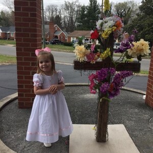 May include: A white dress with short puffed sleeves and a pink ribbon trim at the hem, paired with a pink hair bow. Beside it stands a wooden cross structure adorned with an arrangement of colourful artificial flowers in shades of yellow, purple, and red.