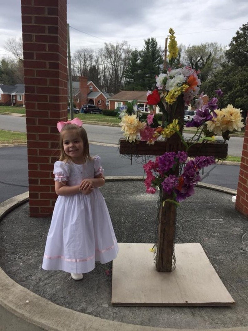 May include: A white dress with short puffed sleeves and a pink ribbon trim at the hem, paired with a pink hair bow. Beside it stands a wooden cross structure adorned with an arrangement of colourful artificial flowers in shades of yellow, purple, and red.