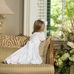 May include: A white dress with short puff sleeves, a round neckline, and a large bow tied at the back waist. The skirt features a subtle pink ribbon trim near the hem. The dress is worn with a white hair bow, showcasing a classic design.