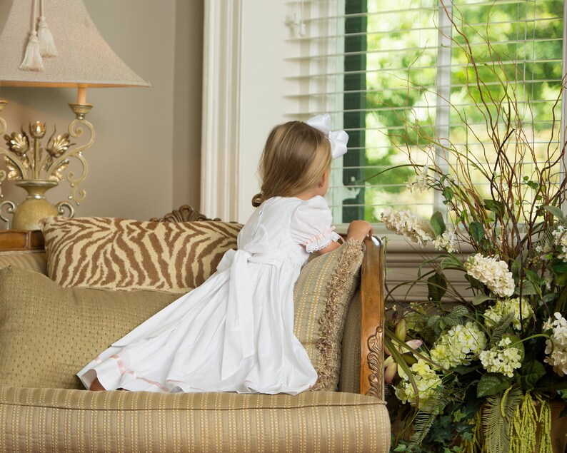 May include: A white dress with short puff sleeves, a round neckline, and a large bow tied at the back waist. The skirt features a subtle pink ribbon trim near the hem. The dress is worn with a white hair bow, showcasing a classic design.