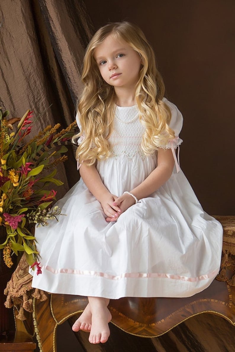 May include: A white dress featuring short puff sleeves with pink ribbon ties, a smocked bodice with delicate embroidery, and a hemline accented by a horizontal pink ribbon. The dress has a round neckline and a full, flowing skirt silhouette.