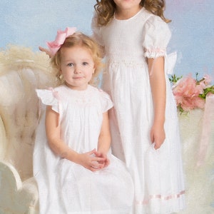 May include: Two white dresses featuring smocked bodices, short puffed sleeves, and light pink ribbon accents. The dresses have a textured fabric, round necklines, and a mid-calf length. Each dress is paired with a matching light pink hair bow.