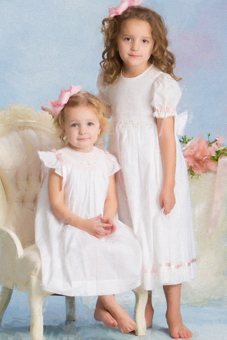 May include: Two white dresses featuring smocked bodices, short puffed sleeves, and light pink ribbon accents. The dresses have a textured fabric, round necklines, and a mid-calf length. Each dress is paired with a matching light pink hair bow.