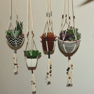 Può includere: Quattro portapiante sospesi in macramè con finte piante grasse. I supporti sono realizzati in corda color crema e presentano perline di legno decorative. I vasi sono di varie forme e colori, tra cui chevron bianco e nero, bianco, marrone e strisce bianche e nere.