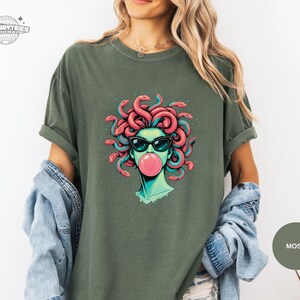 Moss green Comfort Colors tee with the Medusa sunglasses-and-bubblegum design printed front and center. The teal face tones blend well with the muted green shirt for a cool-toned look. Great lifestyle mockup for outdoorsy or casual street style.