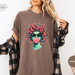 Espresso brown Comfort Colors tee with a centered Medusa portrait wearing black sunglasses and blowing a pink bubblegum bubble. Coral snake hair and teal shading give the graphic a bold pop against the warm neutral tone. Styled for casual winter layering with a flannel vibe.