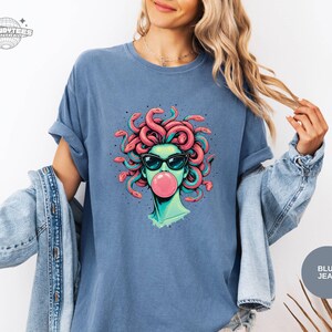 Blue Jean Comfort Colors t-shirt featuring the Medusa portrait graphic with a glossy pink bubblegum bubble. The softer denim-blue color makes the coral snake hair and black sunglasses read crisp and clean. Styled with a light denim jacket for a relaxed weekend outfit.