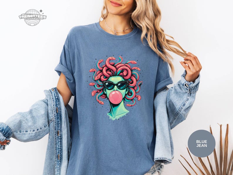 Blue Jean Comfort Colors t-shirt featuring the Medusa portrait graphic with a glossy pink bubblegum bubble. The softer denim-blue color makes the coral snake hair and black sunglasses read crisp and clean. Styled with a light denim jacket for a relaxed weekend outfit.