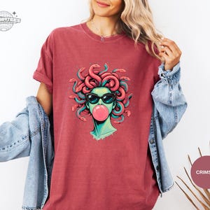 Crimson red Comfort Colors tee with a centered Medusa graphic in sunglasses blowing bubblegum. The coral snake hair echoes the shirt tone while teal accents keep the design from blending in. Strong color option for buyers who want a bolder look.