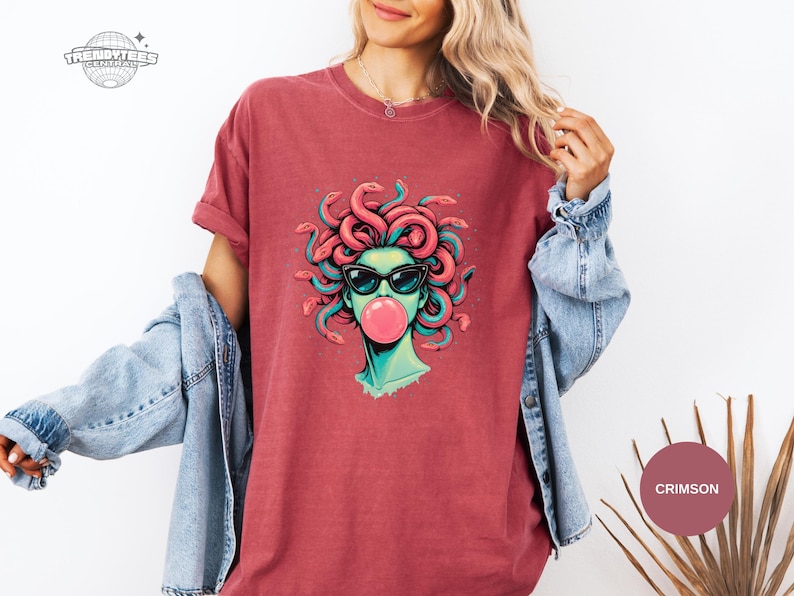 Crimson red Comfort Colors tee with a centered Medusa graphic in sunglasses blowing bubblegum. The coral snake hair echoes the shirt tone while teal accents keep the design from blending in. Strong color option for buyers who want a bolder look.