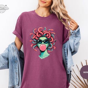 Berry purple Comfort Colors t-shirt showing a bright Medusa bubblegum graphic centered on the chest. The coral snakes and teal highlights stand out strongly on this deeper fashion color. Looks like an easy everyday statement tee for jeans and sneakers.