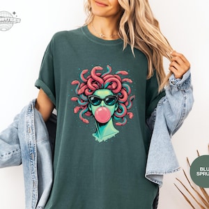 Blue Spruce deep green Comfort Colors t-shirt with the Medusa bubblegum portrait on the front. The dark green background makes the pink bubble and coral snakes pop with high contrast. Styled as a moody, modern mythology aesthetic.