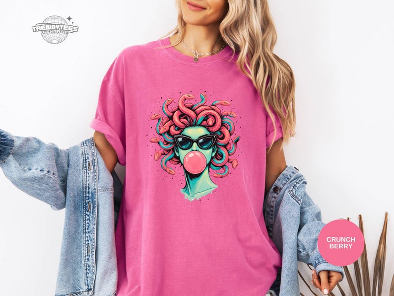 Crunchberry bright pink Comfort Colors tee with the Medusa bubblegum graphic centered. The bold shirt color turns the design into a playful statement while the black sunglasses keep it grounded. This one reads fun, girly, and confident.
