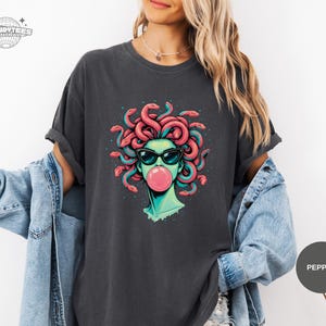Pepper charcoal Comfort Colors tee showing the Medusa graphic centered and sharp. The black sunglasses and darker speck accents feel edgy, while the pink bubble adds a playful twist. This color reads like a versatile, wear-anywhere option.