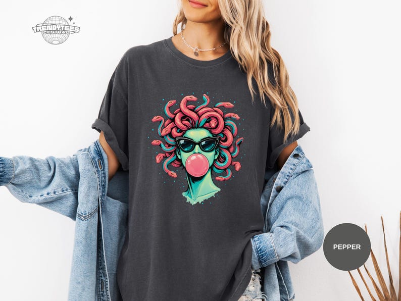 Pepper charcoal Comfort Colors tee showing the Medusa graphic centered and sharp. The black sunglasses and darker speck accents feel edgy, while the pink bubble adds a playful twist. This color reads like a versatile, wear-anywhere option.