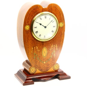 May include: A heart-shaped wooden mantel clock. The clock face has Roman numerals and gold trim. The clock is decorated with inlaid floral and ribbon designs. The base is rectangular and dark brown. The clock measures approximately 25 cm in height.