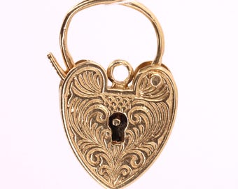 May include: A gold-colored heart-shaped padlock charm featuring an intricate engraved scrollwork pattern across the front surface. The charm includes a central keyhole opening, a small top loop for attachment, and a hinged curved bail at the top.
