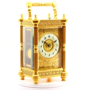 Puede incluir: Un reloj antiguo de color dorado adornado, con una esfera de vidrio y números romanos. El reloj tiene una manija decorativa en la parte superior y se asienta sobre una base blanca.