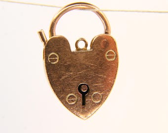 May include: A heart-shaped gold padlock with a keyhole and a curved handle. The padlock is suspended against a white background. The lock has a vintage or antique appearance, with visible screw heads.