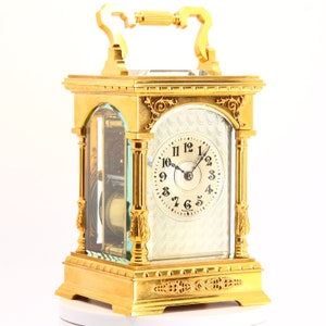 Puede incluir: Un reloj de mesa de color dorado, adornado, con una caja de cristal y una esfera blanca. El reloj tiene una esfera con números romanos y una aguja negra para las horas y los minutos. El reloj está sobre una superficie blanca.