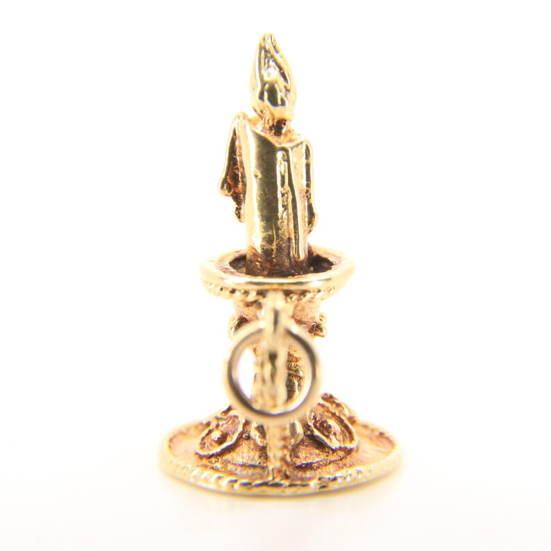 Puede incluir: Un charm de vela dorado con un dise&ntilde;o detallado. El charm presenta una vela en un soporte con una base redonda y un lazo para sujetarlo a una pulsera o collar. La vela tiene una llama en la parte superior.