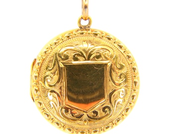 May include: A gold locket pendant with an ornate design featuring a shield in the center. The locket is round and has a detailed border.