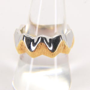Puede incluir: Un anillo bicolor con un diseño único. La parte superior es de color plateado, mientras que la sección inferior presenta una banda texturizada de color dorado. El anillo se exhibe sobre un soporte transparente, mostrando sus intrincados detalles.