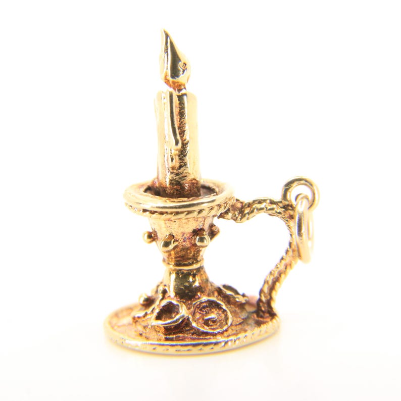 Puede incluir: Un charm de vela dorado con un dise&ntilde;o detallado. El charm presenta una vela con una llama, colocada en un candelabro decorativo con un asa. El charm probablemente est&aacute; hecho de oro y est&aacute; dise&ntilde;ado para ser adjuntado a una pulsera o collar.