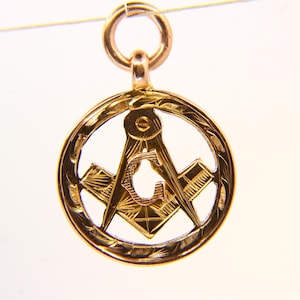 May include: A gold-colored pendant featuring the Masonic Square and Compasses symbol within a circular frame. The pendant has a detailed, engraved design and a small loop at the top for a chain. The pendant is suspended against a white background.
