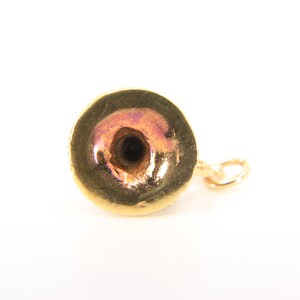 Puede incluir: Un peque&ntilde;o dije de color dorado con forma circular y un agujero central. El dije tiene un brillo met&aacute;lico con toques de rosa y morado. Un peque&ntilde;o cierre dorado est&aacute; unido al costado.