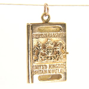 Könnte beinhalten: Ein goldfarbener Metallanhänger in Form eines Reisepasses. Das Vordercover zeigt den geprägten Text BRITISH PASSPORT und UNITED KINGDOM BRITAIN N IRELAND sowie ein zentrales Wappenemblem. Ein kleiner Ring ist an der Oberseite befestigt.