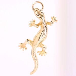 May include: A gold-toned metal pendant shaped like a lizard with a textured surface and detailed limbs. The lizard is depicted in a climbing position with its body curved and a small circular jump ring attached to the top of its head for hanging.