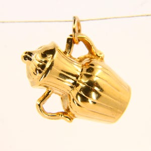 May include: A gold-colored charm in the shape of a pitcher or vase. The charm has a detailed design and is made of metal.