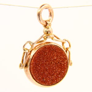 May include: A gold-toned fob chain with a round, red-brown stone pendant. The pendant is set in a gold-toned frame and has a textured surface.